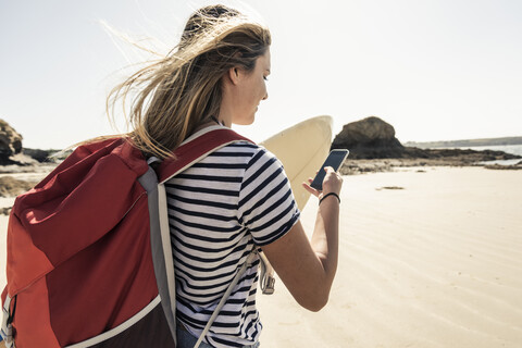 Junge Frau am Strand, mit Surfbrett, mit Smartphone, lizenzfreies Stockfoto