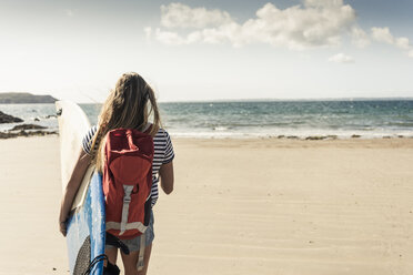 Junge Frau mit Rucksack, die am Strand spazieren geht und ein Surfbrett trägt - UUF16435