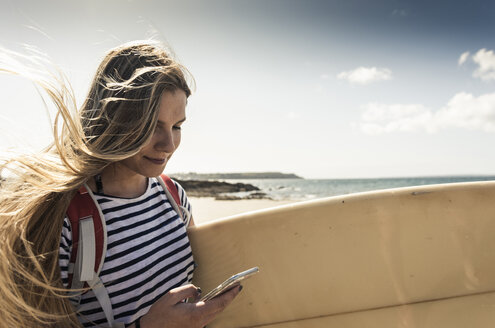 Junge Frau am Strand, mit Surfbrett, mit Smartphone - UUF16436