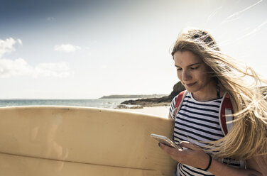 Junge Frau am Strand, mit Surfbrett, mit Smartphone - UUF16437