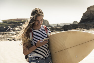 Junge Frau am Strand, mit Surfbrett, mit Smartphone - UUF16438
