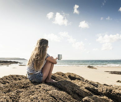 Junge Frau sitzt auf einem Felsen am Strand und entspannt sich mit einer Tasse Tee - UUF16446