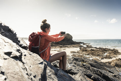 Junge Frau sitzt an einem felsigen Strand und benutzt ihr Smartphone - UUF16453