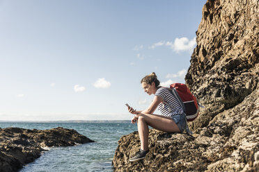 Young woman sitting on a rocky beach, using smartphone - UUF16463