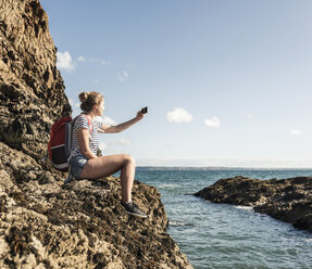 Young woman sitting on a rocky beach, taking pictures with her smartphone - UUF16464