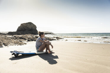 Junge Frau sitzt auf ihrem Surfbrett am Strand und schaut aufs Meer - UUF16468