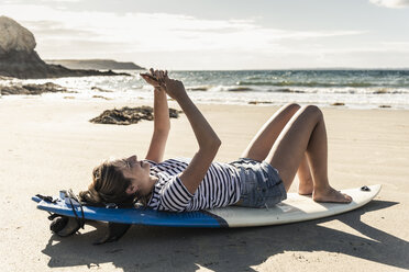 Junge Frau am Strand, entspannt auf dem Surfbrett, mit Smartphone - UUF16475