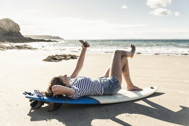 Junge Frau am Strand, entspannt auf dem Surfbrett, mit Smartphone - UUF16476