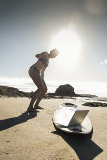 Young woman standing by surfboard, using laptop - UUF16480