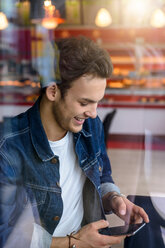 Männlicher Student, der in einem Café am Fensterplatz auf sein Smartphone schaut, Blick durch das Fenster - CUF47139