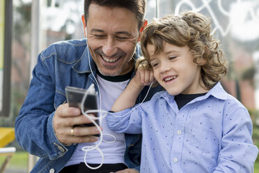 Happy father and son sharing cell phone and earbuds - MAUF02274