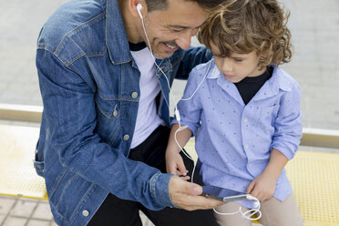 Father and son sitting at tram stop in the city sharing cell phone and earbuds - MAUF02275