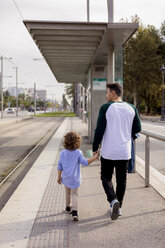 Rückansicht von Vater und Sohn, die Hand in Hand an einer Straßenbahnhaltestelle in der Stadt gehen - MAUF02279