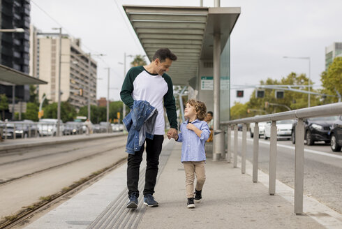 Lächelnder Vater und Sohn gehen Hand in Hand an einer Straßenbahnhaltestelle in der Stadt - MAUF02289