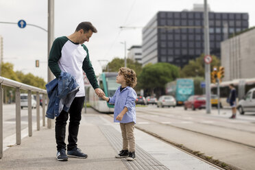 Lächelnder Vater und Sohn stehen Hand in Hand an einer Straßenbahnhaltestelle in der Stadt - MAUF02290