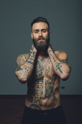 Portrait of young man with beard, bare chest covered in tattoos, hands behind neck - CUF47205