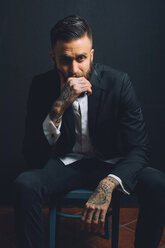 Portrait of young man, sitting on chair, wearing suit, tattoos on hands, pensive expression - CUF47215