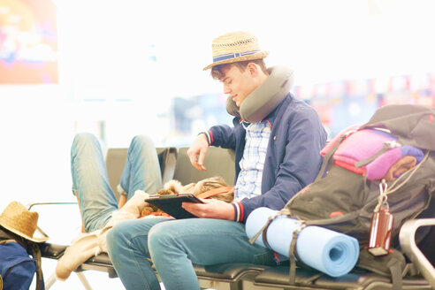 Junges Paar in der Abflughalle eines Flughafens, die Frau stützt den Kopf auf die Beine des Mannes, der Mann benutzt ein digitales Tablet - CUF47305