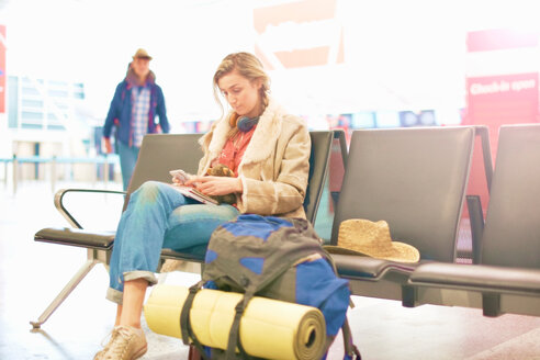 Junge Frau auf dem Flughafen, sitzend mit Rucksack neben sich, mit Smartphone - CUF47306