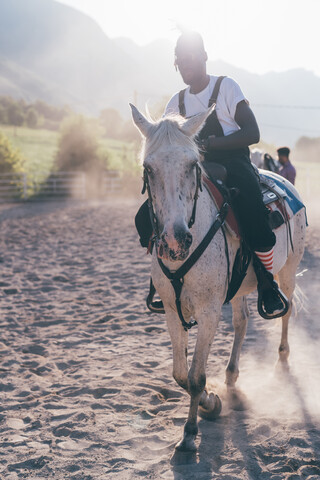 Junger Hipster-Mann reitet auf einem Pferd in einer sonnenbeschienenen Reithalle, Primaluna, Trentino-Südtirol, Italien, lizenzfreies Stockfoto