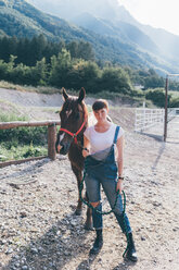 Junge Frau, die ein Pferd in einer ländlichen Reithalle führt, Porträt in voller Länge - CUF47531