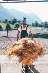 Cool young man with wheelbarrow hay in rural equestrian arena, portrait - CUF47534