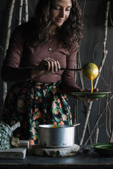Junge Frau schöpft frische Suppe aus einem Kochtopf an einer rustikalen Küchentheke - CUF47548
