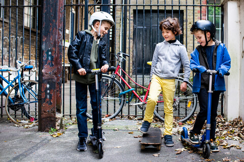 Jungen fahren Tretroller und Skateboard, Fahrräder im Hintergrund, lizenzfreies Stockfoto