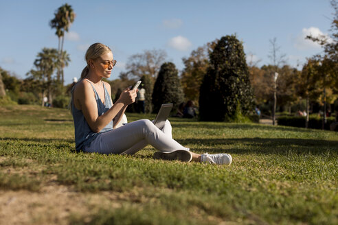 Junge Frau sitzt im Park und benutzt Handy und Laptop - MAUF02298