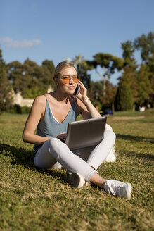 Junge Frau sitzt im Park und benutzt Handy und Laptop - MAUF02299