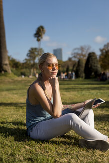 Junge Frau sitzt im Park mit Handy und Laptop - MAUF02300