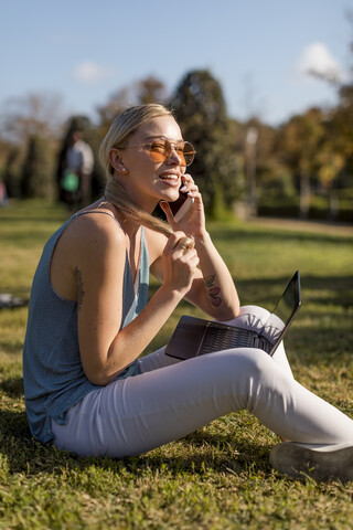 Junge Frau sitzt im Park und benutzt Handy und Laptop, lizenzfreies Stockfoto