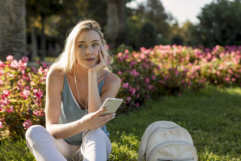 Lächelnde junge Frau sitzt im Park mit Handy und Ohrhörern, lizenzfreies Stockfoto