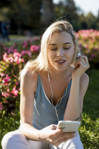 Lächelnde junge Frau sitzt im Park mit Handy und Ohrhörern, lizenzfreies Stockfoto