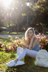 Portrait of smiling young woman sitting in park with cell phone and earbuds - MAUF02308