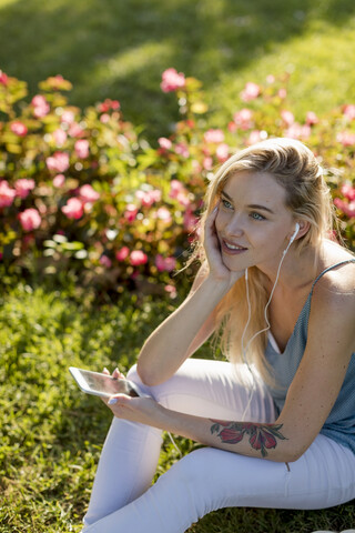 Smiling young woman sitting in park with cell phone and earbuds stock photo