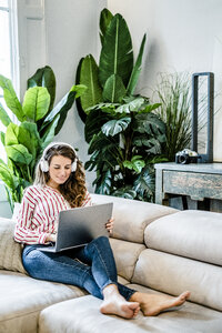 Lächelnde Frau mit Laptop und Kopfhörern auf der Couch sitzend - GIOF05513