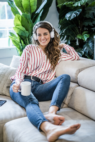Porträt einer lächelnden Frau mit einer Tasse Kaffee und Kopfhörern auf der Couch sitzend, lizenzfreies Stockfoto