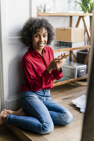 Porträt einer lächelnden Frau, die mit ihrem Handy zu Hause auf dem Boden sitzt, lizenzfreies Stockfoto