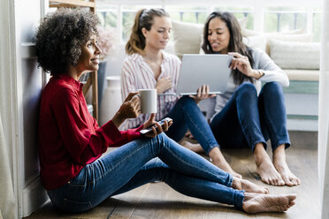 Three women with cell phone, coffee cup and laptop sitting on the floor at home - GIOF05525