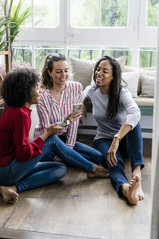 Drei glückliche Frauen, die zu Hause mit ihren Handys auf dem Boden sitzen, lizenzfreies Stockfoto