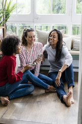 Three happy women sitting on the floor at home with cell phones - GIOF05526