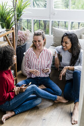Three happy women sitting on the floor at home with cell phones - GIOF05527