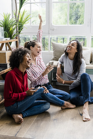 Drei unbekümmerte Frauen, die zu Hause mit ihren Handys auf dem Boden sitzen, lizenzfreies Stockfoto