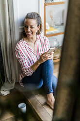 Laughing woman sitting on the floor at home with cell phone - GIOF05531