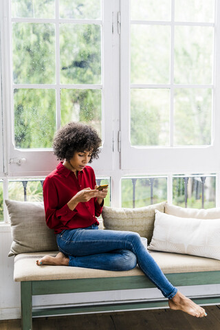 Ernste Frau, die zu Hause am Fenster sitzt und ein Mobiltelefon benutzt, lizenzfreies Stockfoto