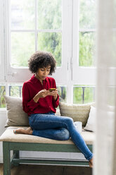 Serious woman sitting at the window at home using cell phone - GIOF05535