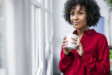 Lächelnde Frau mit einer Tasse Kaffee, die zu Hause aus dem Fenster schaut - GIOF05538