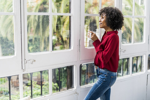 Lächelnde Frau mit einer Tasse Kaffee, die zu Hause aus dem Fenster schaut, lizenzfreies Stockfoto