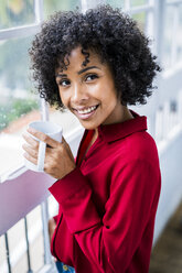 Portrait of smiling woman with cup of coffee at the window at home - GIOF05541